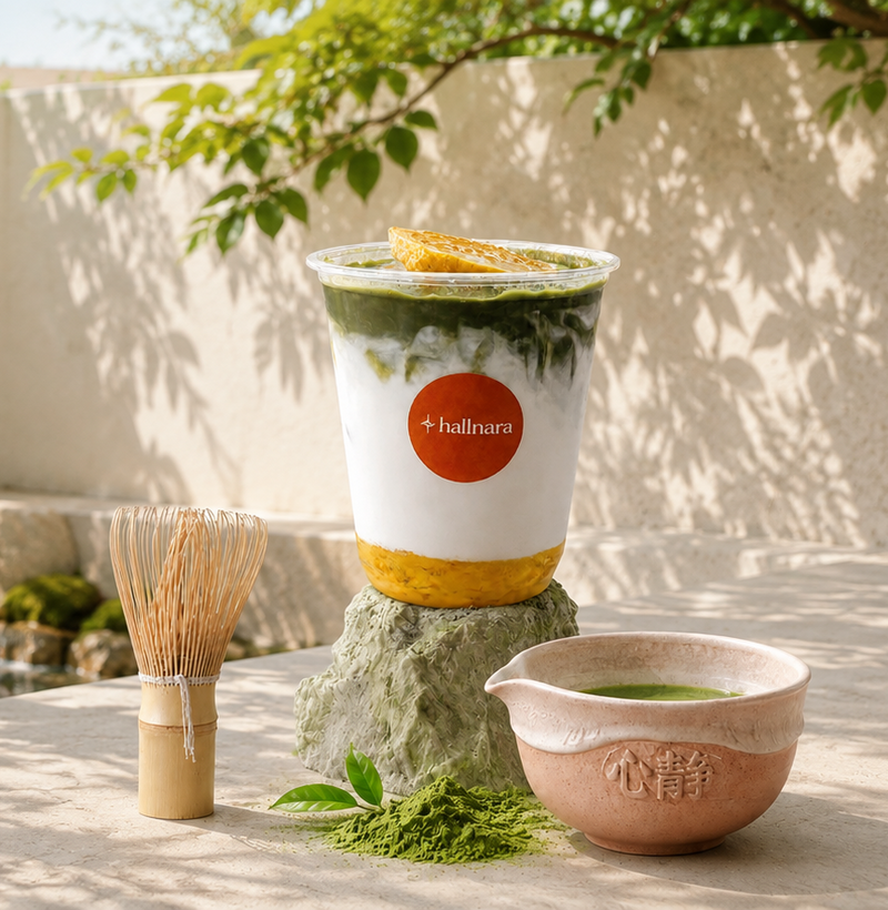 A layered matcha green tea drink in a clear plastic cup with a red circular +ballmara logo, set on a stone pedestal outdoors. A bamboo whisk, green tea powder, and a small ceramic bowl are arranged around it on a stone surface with leafy branches and a beige wall in the background.