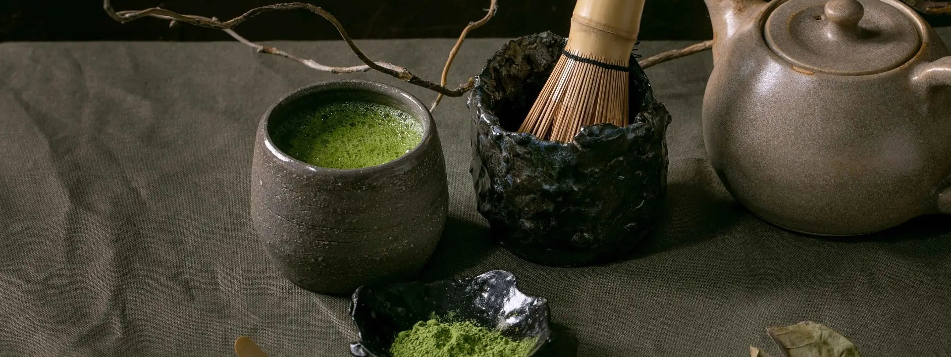 Tea-making setup with a cup of green tea, whisk, and teapot on a textured surface.