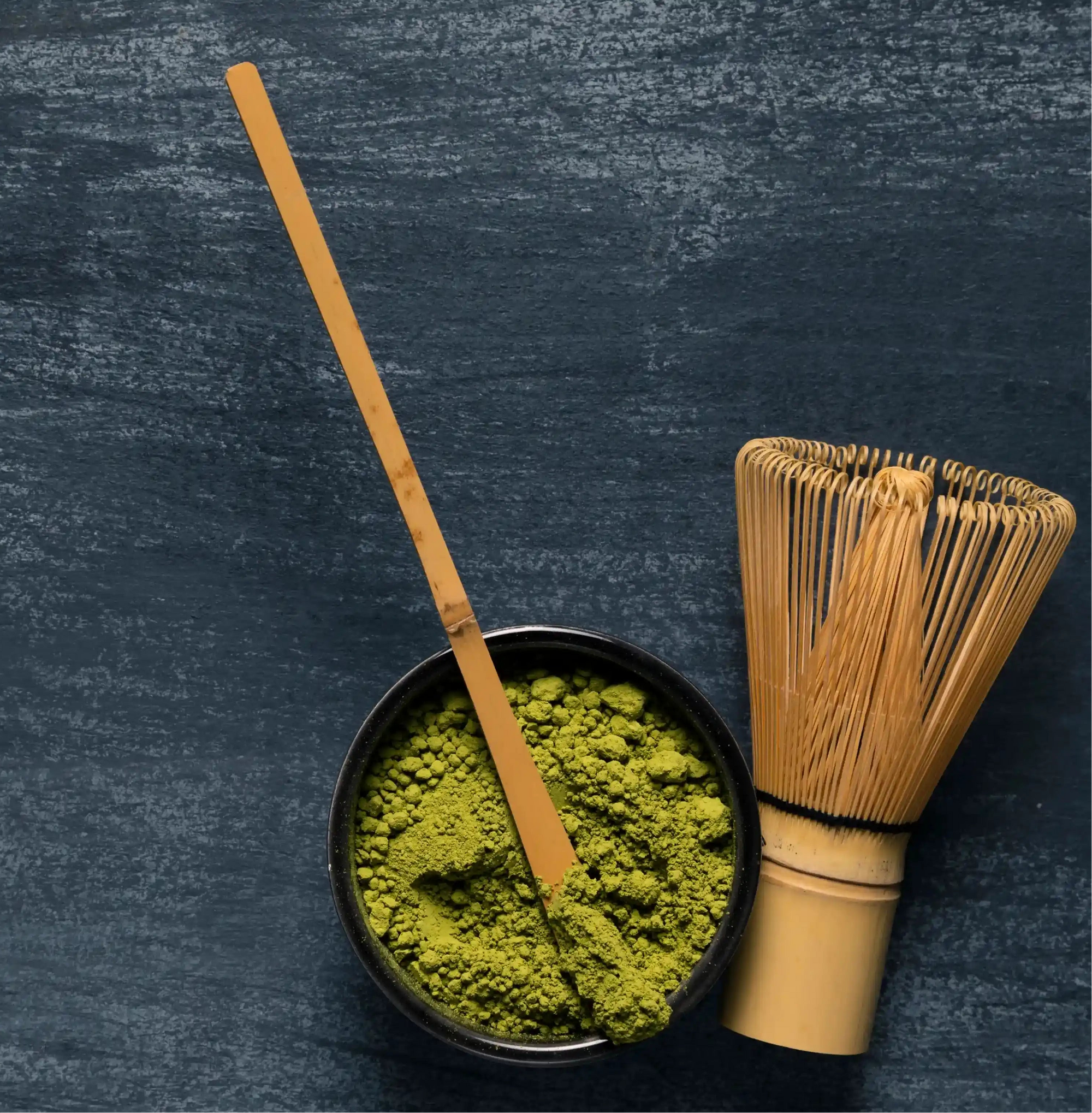 Matcha green tea powder in a black bowl with a wooden whisk on a dark textured background